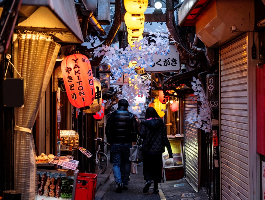 Ruelle japonaise traditionnelle avec des lanternes rouges et jaunes éclairées, des enseignes en japonais, des branches décoratives blanches, et deux personnes marchant au centre, dans une ambiance chaleureuse et conviviale