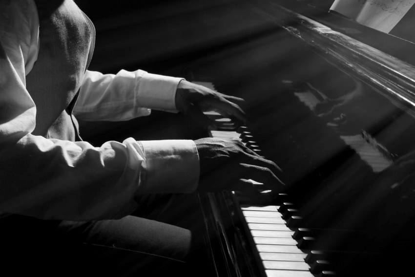 Homme jouant du piano en noir et blanc, éclairé par des rayons de lumière, illustrant l'apprentissage et la pratique de la musique.